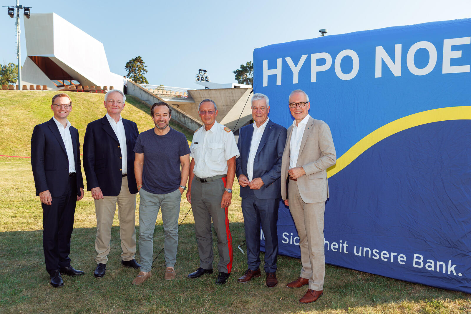 sechs Männer in Anzug stehen vor einem blauen Würfel mit Schriftzug "HYPO NOE" auf einer Wiese