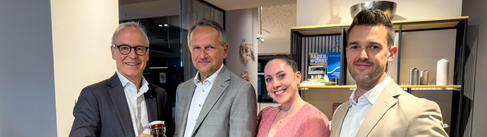 Drei Männer im Anzug und eine Frau im Kleid stehen beisammen. Ein Mann überreicht einem anderen eine Flasche Bier.