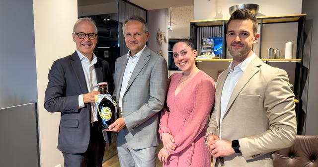 Drei Männer im Anzug und eine Frau im Kleid stehen beisammen. Ein Mann überreicht einem anderen eine Flasche Bier.