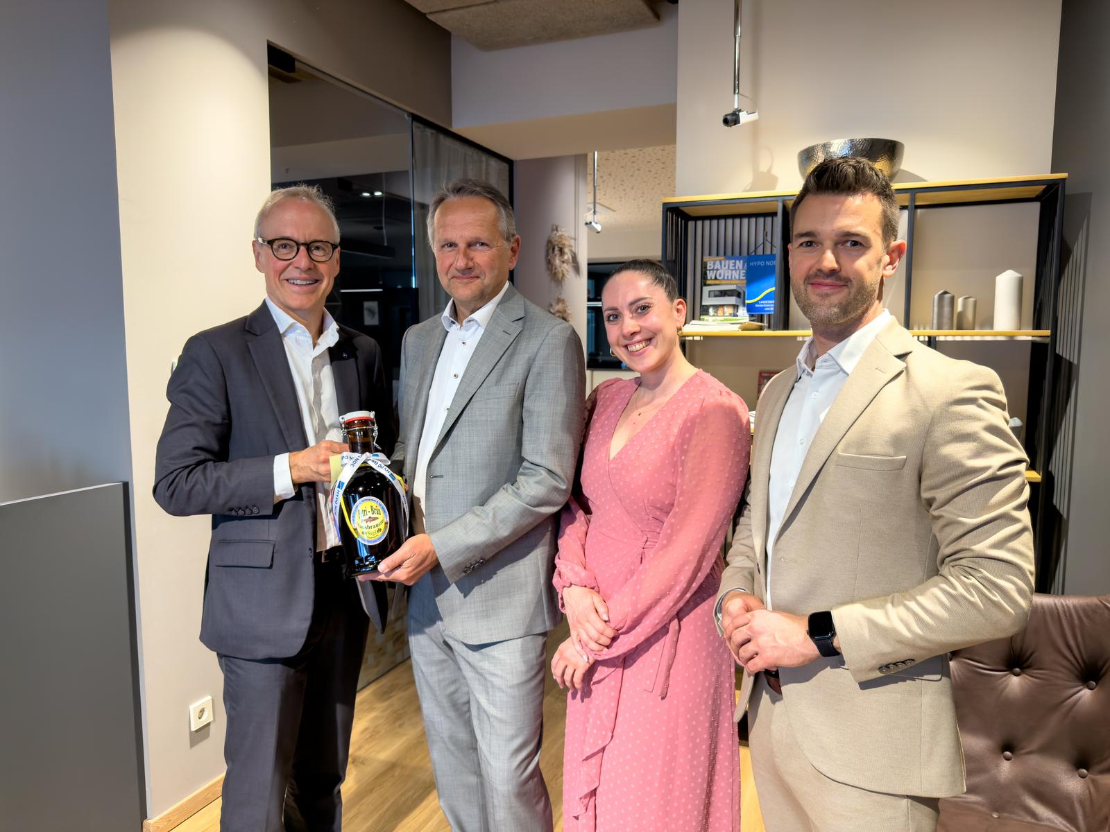 Drei Männer im Anzug und eine Frau im Kleid stehen beisammen. Ein Mann überreicht einem anderen eine Flasche Bier.
