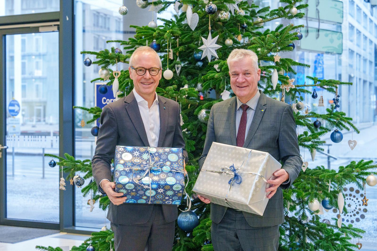 Zwei Männer in Anzug stehen vor einem geschmückten Christbaum und halten je ein Geschenkspackerl in der Hand.
