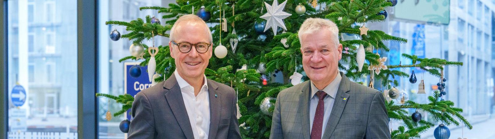 Zwei Männer in Anzug stehen vor einem geschmückten Christbaum und halten je ein Geschenkspackerl in der Hand.