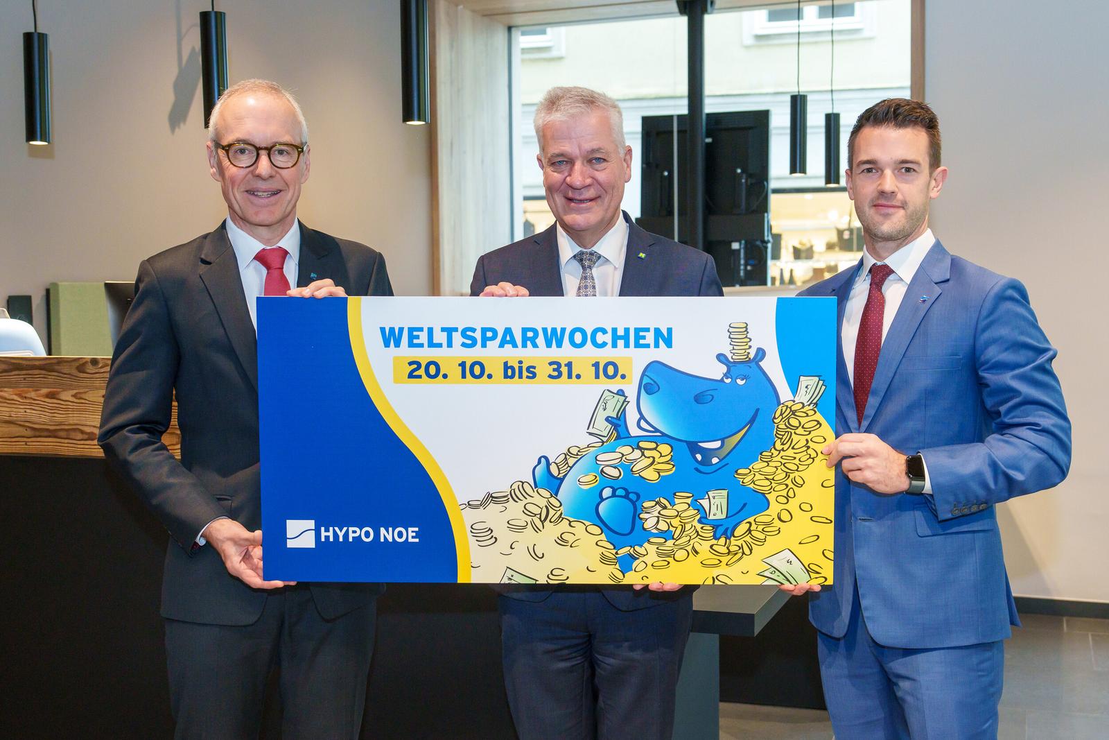 Drei Männer in Anzug und Krawatte halten ein Schild mit einem aufgedruckten Hippo und der Aufschrift "HYPO NOE Weltsparwochen von 20.10 bis 31.10.".