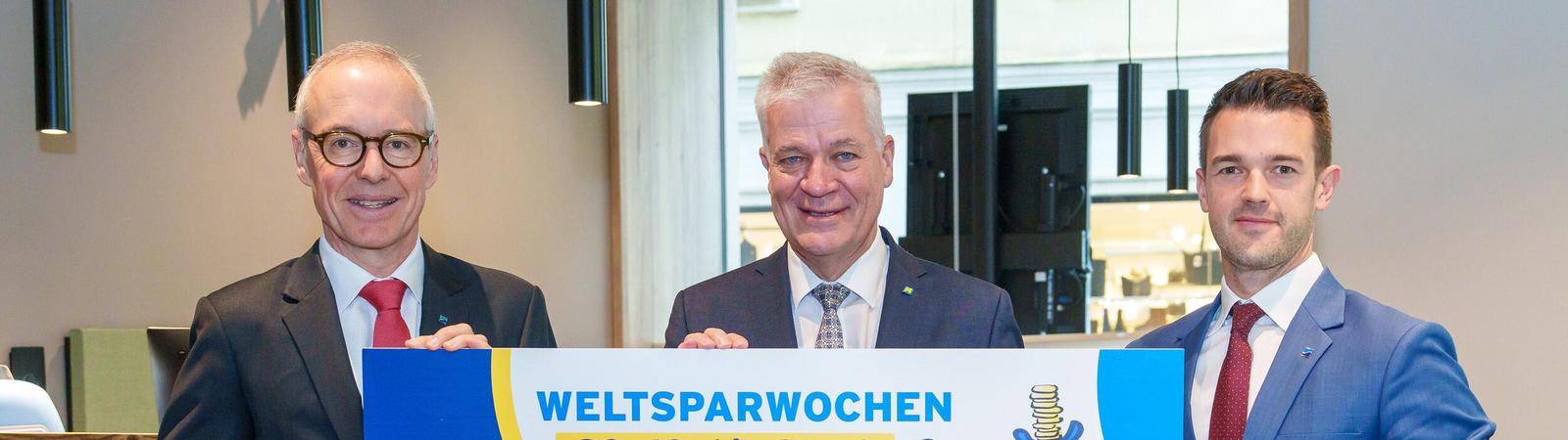 Drei Männer in Anzug und Krawatte halten ein Schild mit einem aufgedruckten Hippo und der Aufschrift "HYPO NOE Weltsparwochen von 20.10 bis 31.10.".