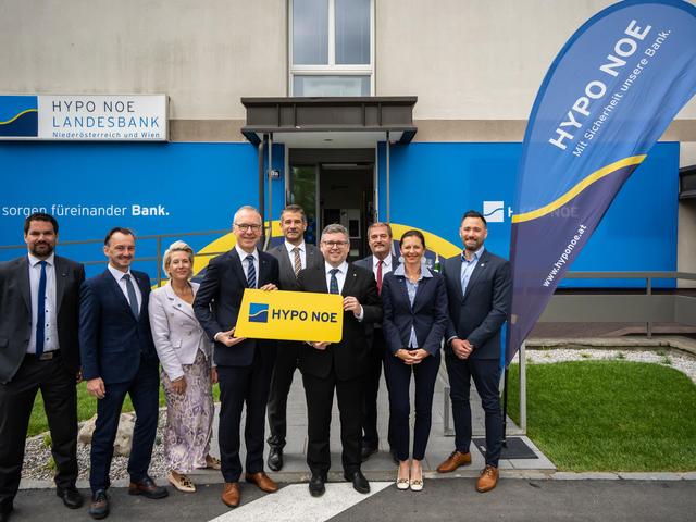 Eine Gruppe von Menschen in Business-Kleidung steht vor einem Gebäude, dessen Fassade mit dem blau-gelben Logo der HYPO NOE versehen ist.