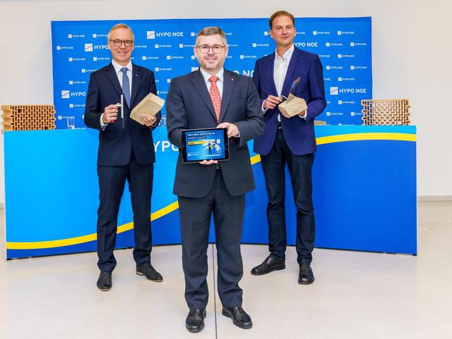 Drei Männer in Anzug und Krawatte, zwei halten Ziegelsteine und Maurerkelle, einer ein Tablet mit einer Grafik.