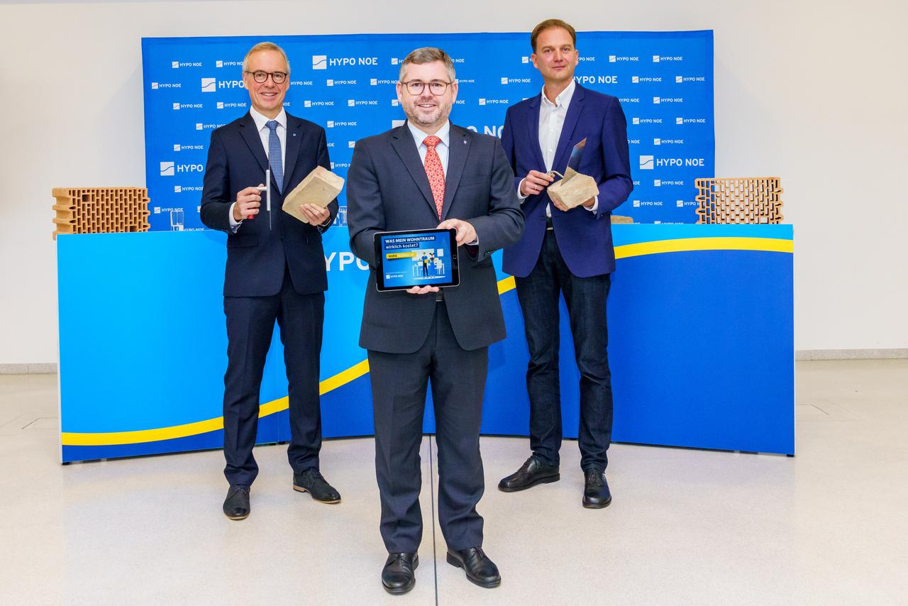 Drei Männer in Anzug und Krawatte, zwei halten Ziegelsteine und Maurerkelle, einer ein Tablet mit einer Grafik.