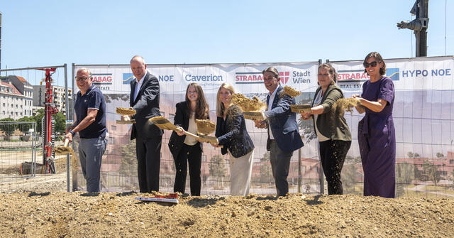 Sieben Menschen in Business Outfit stehen auf einer Baustelle und halten je einen Spaten in der Hand.
