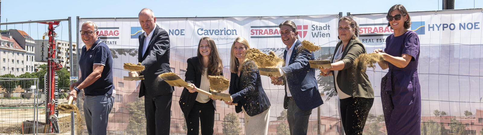 Sieben Menschen in Business Outfit stehen auf einer Baustelle und halten je einen Spaten in der Hand.