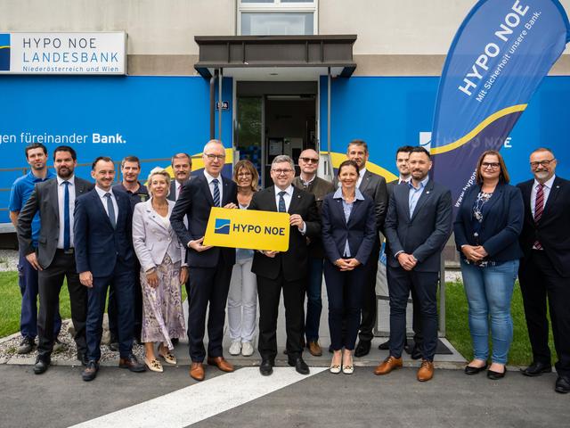Eine Gruppe von Menschen in Business-Kleidung steht vor einem Gebäude, dessen Fassade mit dem blau-gelben Logo der HYPO NOE versehen ist.