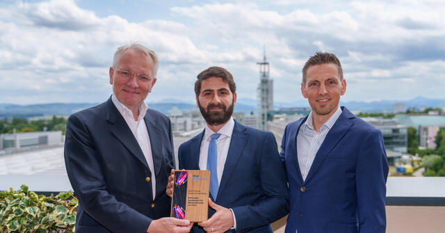 Drei Männer in Anzug halten einen Award in die Kamera, rundherum sind Pflanzen und im Hintergrund eine Kirche zu sehen.
