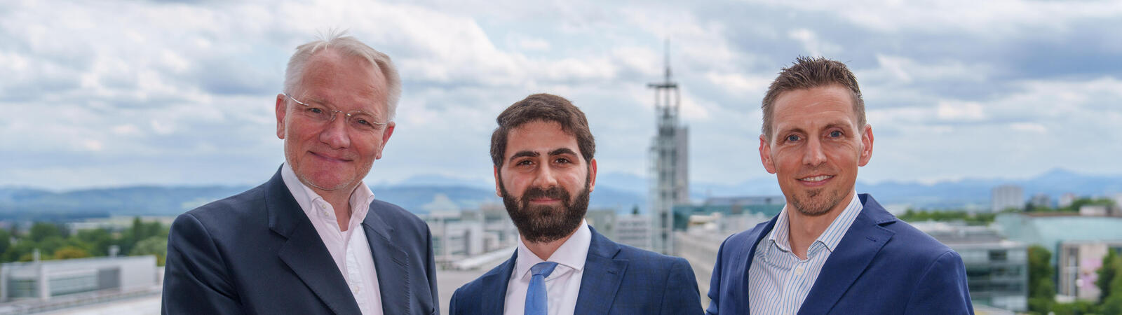 Drei Männer in Anzug halten einen Award in die Kamera, rundherum sind Pflanzen und im Hintergrund eine Kirche zu sehen.