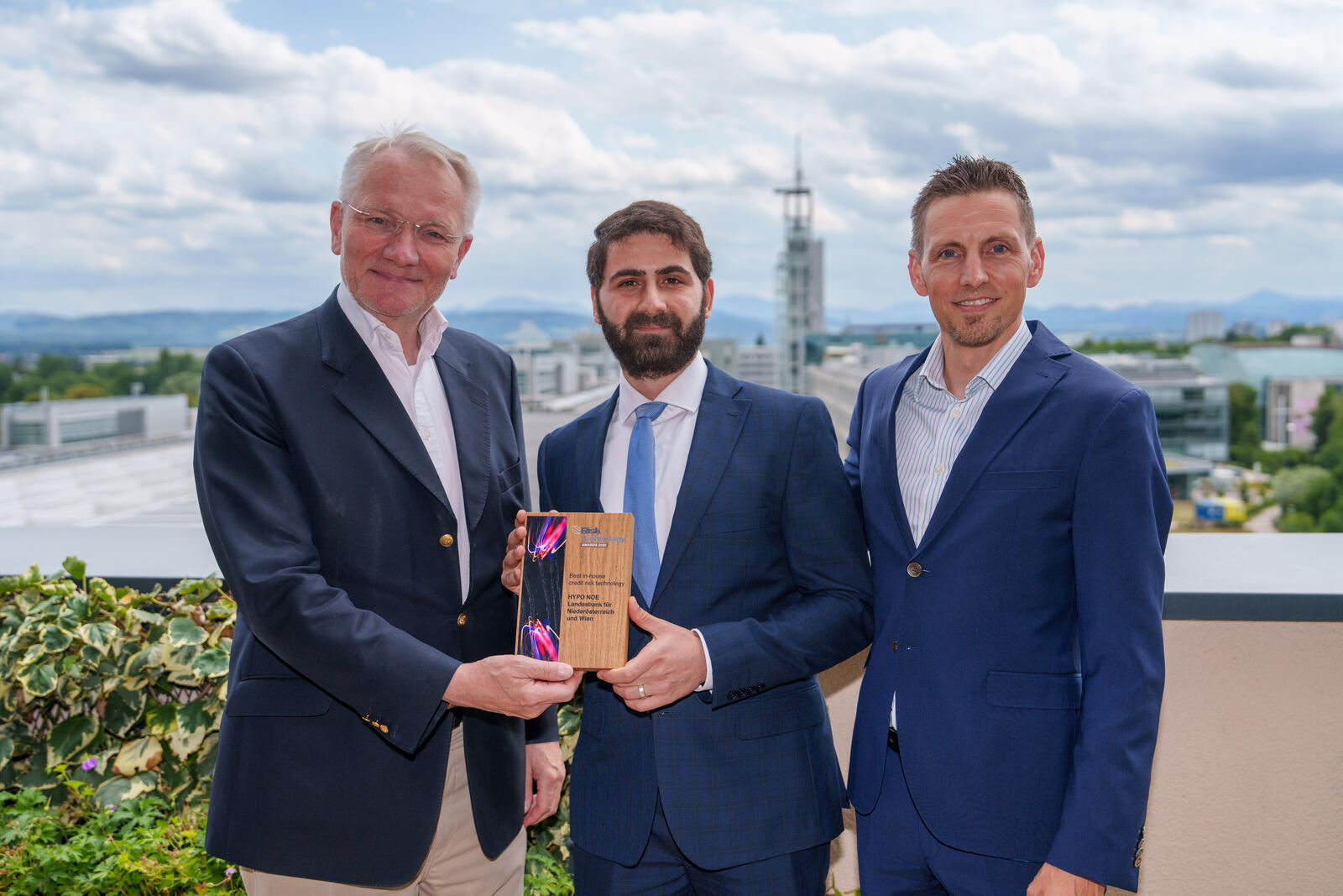 Drei Männer in Anzug halten einen Award in die Kamera, rundherum sind Pflanzen und im Hintergrund eine Kirche zu sehen.