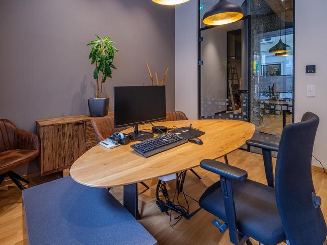 ein Schreibtisch mit Bildschirm und Bürosessel, im Hintergrund ein niedriges Regal mit Pflanze.