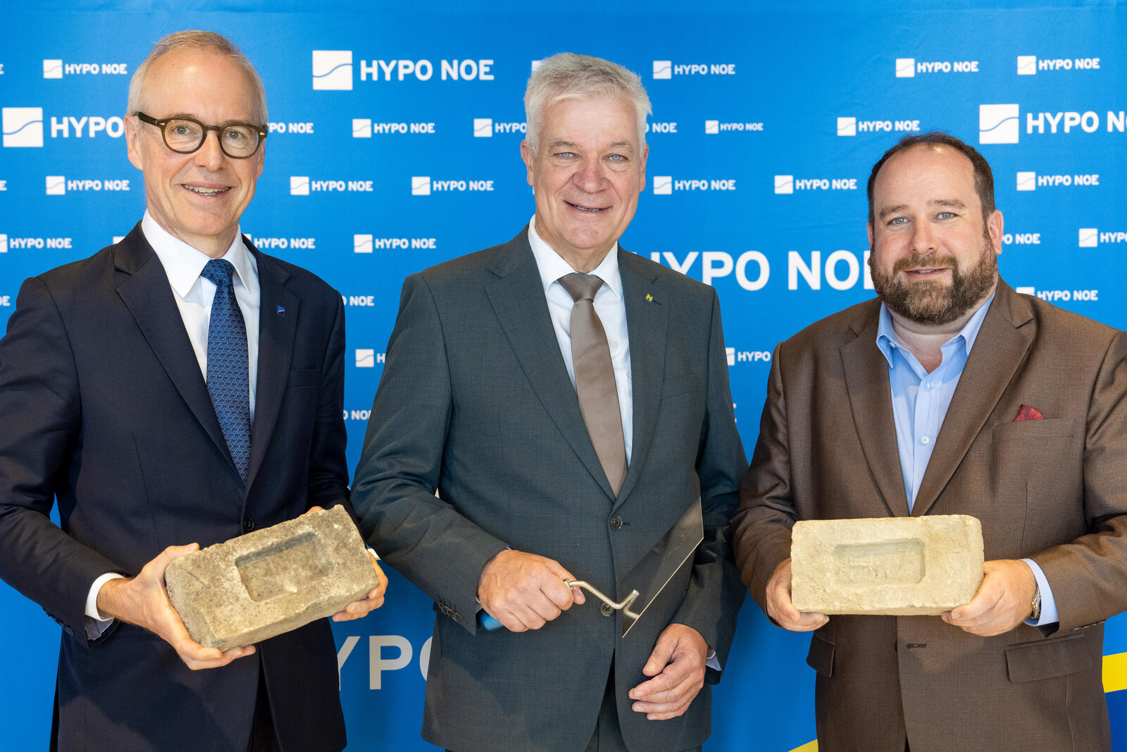 Drei Männer in Anzug und Krawatte präsentieren Ziegelsteine.