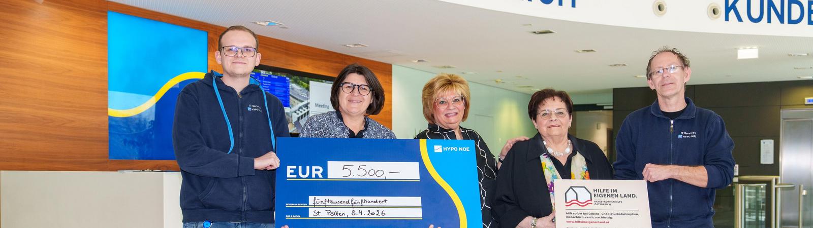 Zwei Männer und drei Frauen halten zwei Schilder: Einen Spendenscheck in blau mit 5.500 Euro und ein Schild mit Vereinslogo von Hilfe im eigenen Land.