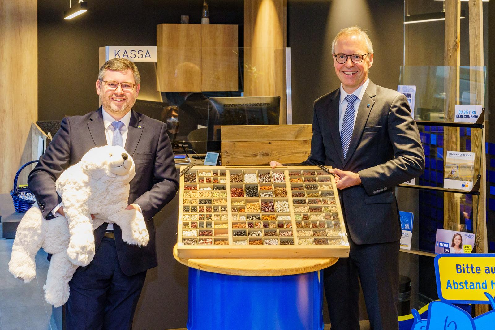 Zwei Männern in Anzug und Krawatte, einer hält einen Stoffeisbären, ein anderer hält einen großen Samenkasten