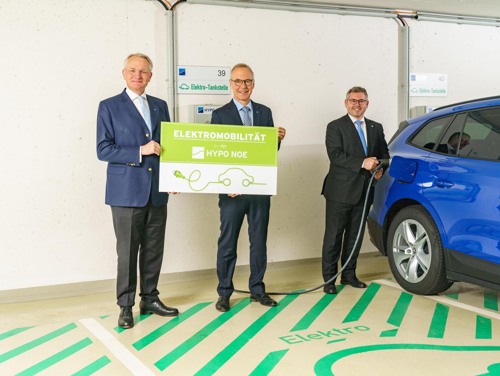 Drei Männer in Anzug und Krawatte stehen in einer Garage vor einer E-Ladesäule und einem blauen Auto und zwei halten ein Schild, einer steckt Auto an.