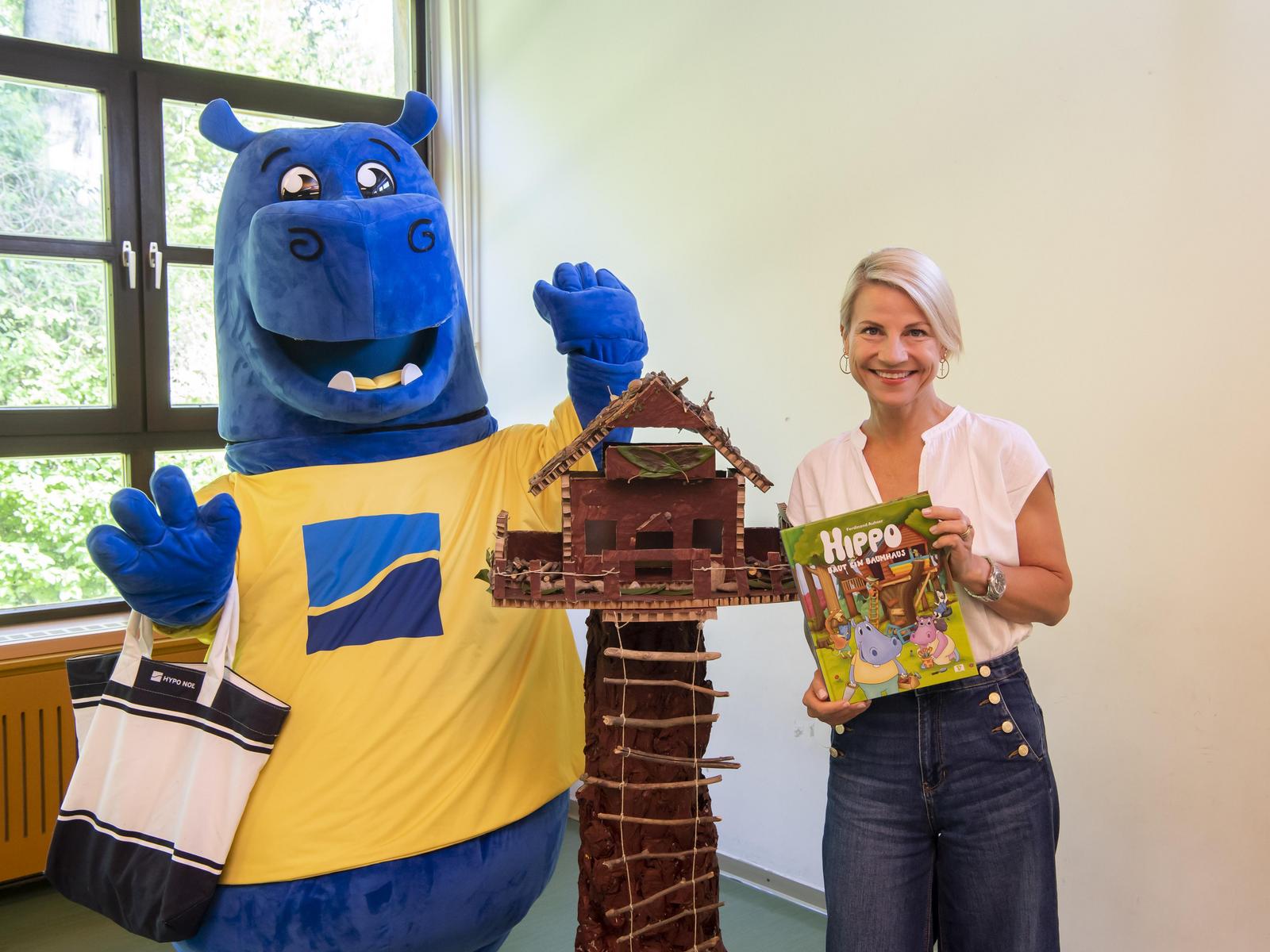 Hippo-Maskottchen mit dem Modell eines Baumhauses und einer Frau, die ein Kinderbuch hält.