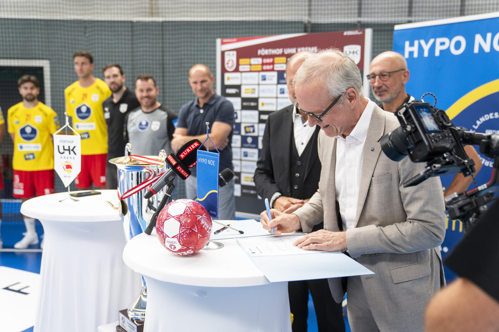 Unterzeichnung einer Sponsoringvereinbarung, dahinter Handballteam und Medien.