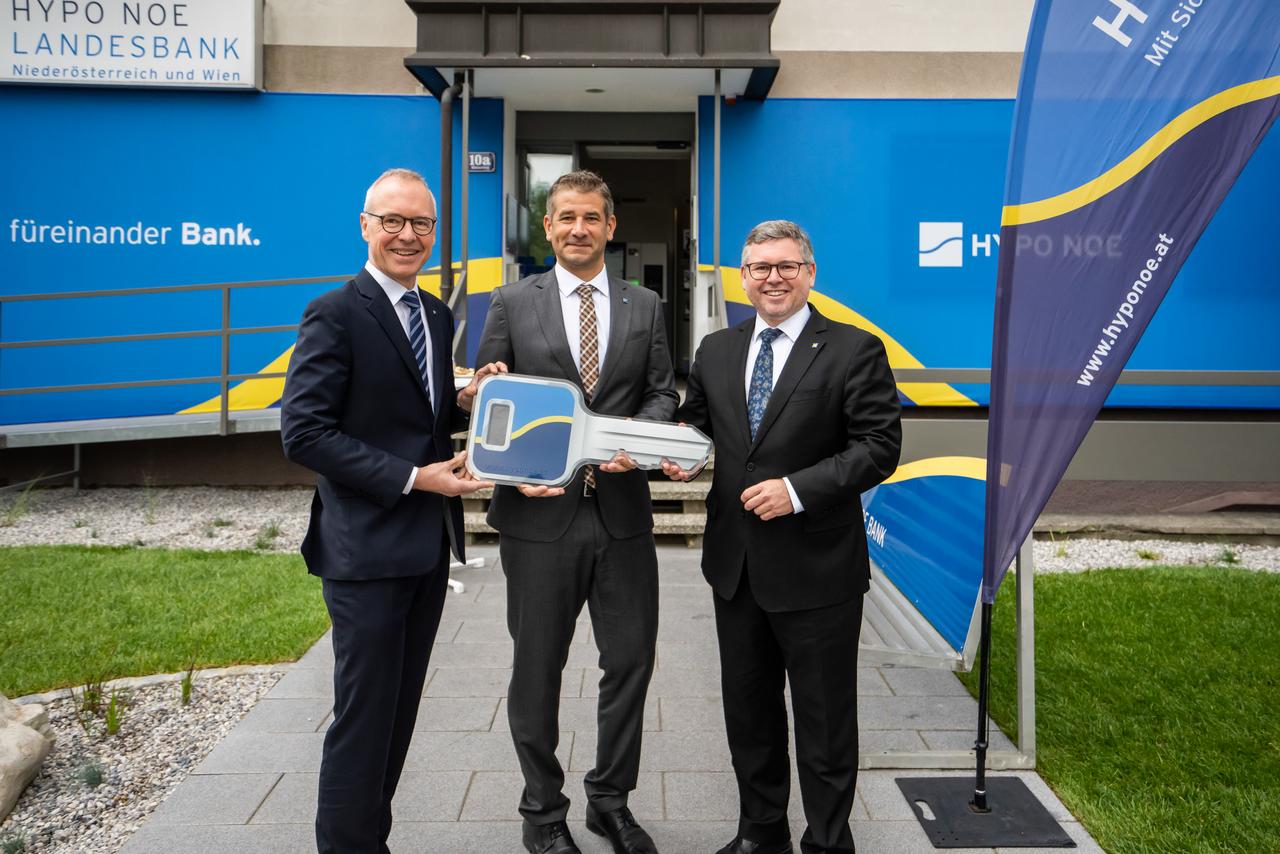 Drei Männer mittleren Alters in Anzügen stehen vor einem Gebäude, dessen Fassade mit dem blau-gelben Logo der HYPO NOE versehen ist.