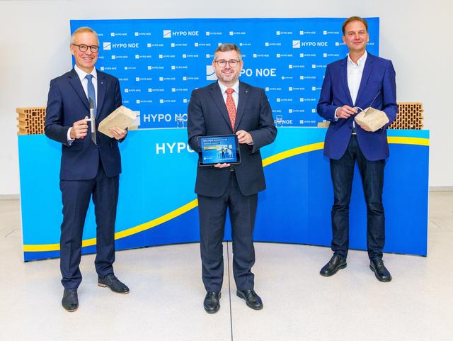 Drei Männer in Anzug und Krawatte, zwei halten Ziegelsteine und Maurerkelle, einer ein Tablet mit einer Grafik.