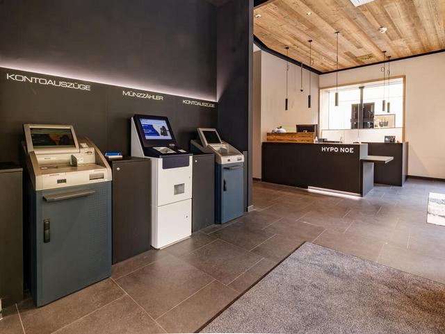 Drei Bank-Automaten links im Vordergrund vor einer anthrazitfarbenen Wand, rechts im Hintergrund ist der Bankschalter zu sehen.