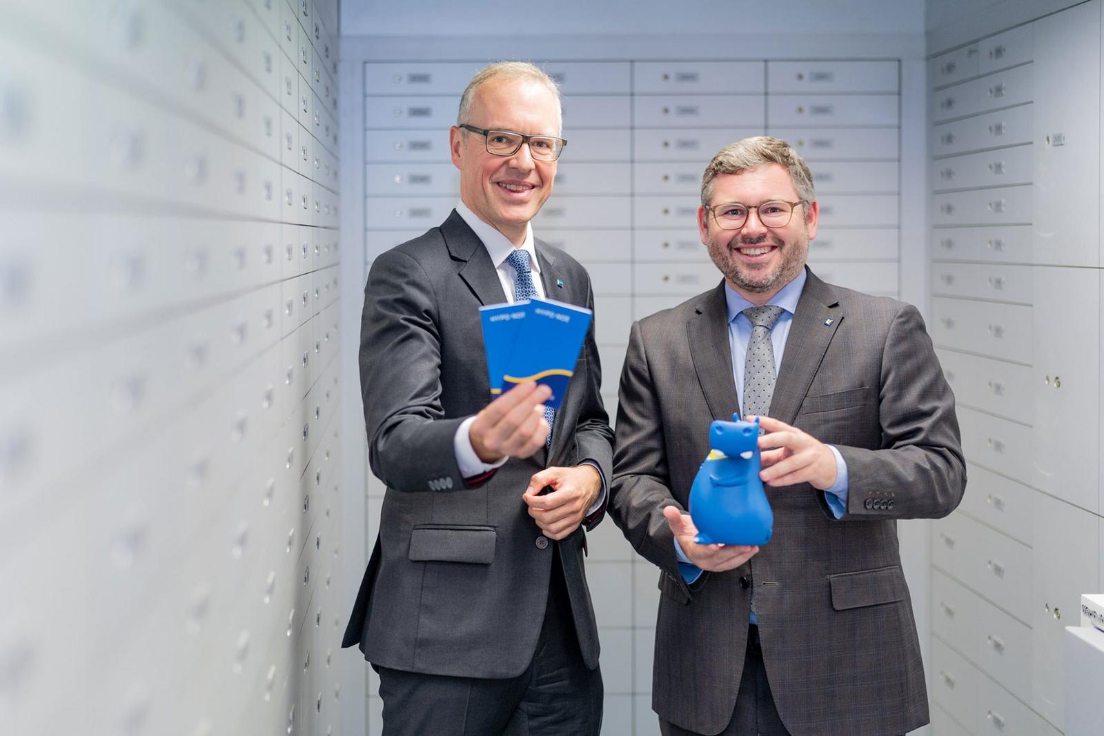 zwei Männer mittleren Alters in Anzug und Krawatte, einer hält zwei Sparbücher, der andere eine Spardose in Form eines Flusspferdes
