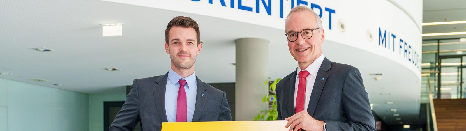 Zwei Männer mittleren Alters im Anzug mit Krawatte stehen im Eingangsbereich eines Bürogebäudes und halten ein Schild.