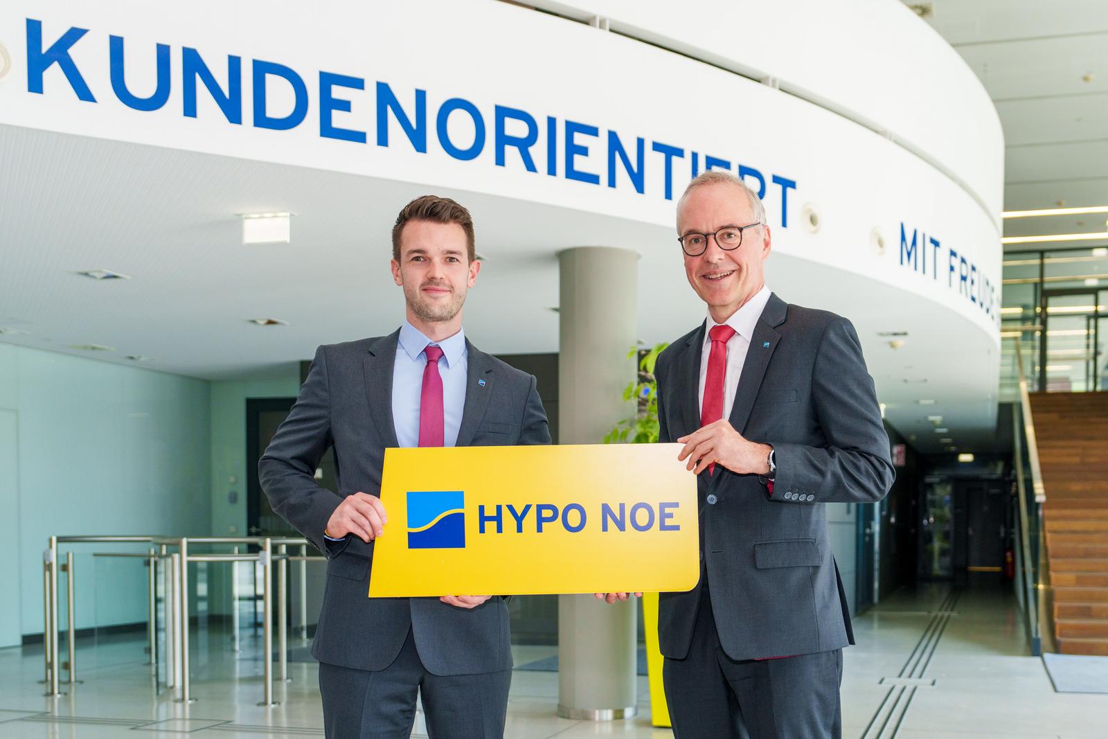 Zwei Männer mittleren Alters im Anzug mit Krawatte stehen im Eingangsbereich eines Bürogebäudes und halten ein Schild.