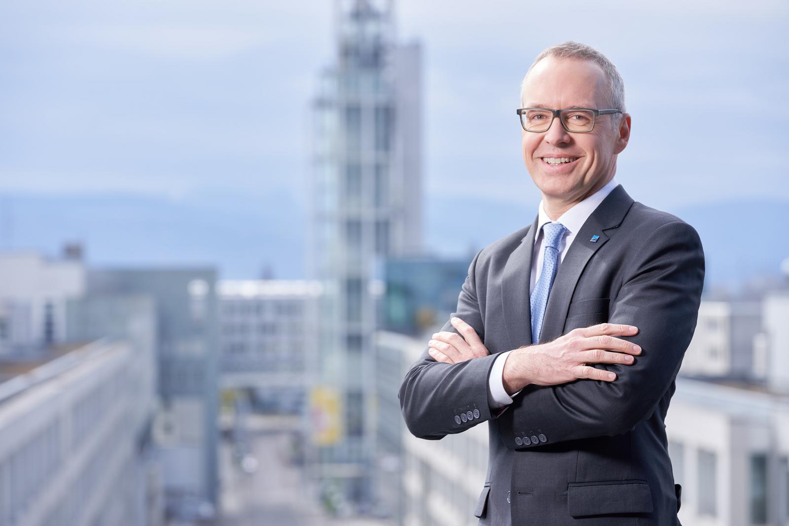 Portrait eines Mannes mittleren Alters in Anzug und Krawatte, im Hintergrund ist ein Turm und Gebäude zu sehen.