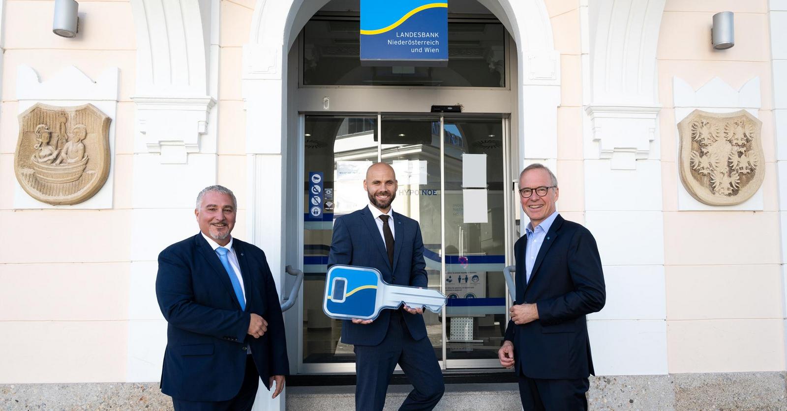 Drei Männer in Anzug und Krawatte stehen vor dem Eingang zur HYPO NOE Filiale, einer hält einen übergroßen Schlüssel.
