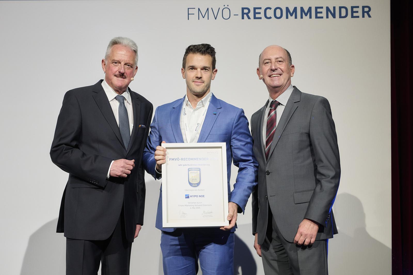 Drei Männer in Anzügen vor einem weißen Hintergrund, der mittlere Mann hält eine Urkunde in der Hand.