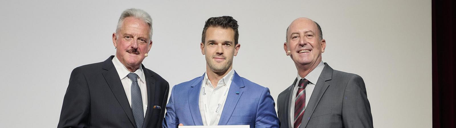 Drei Männer in Anzügen vor einem weißen Hintergrund, der mittlere Mann hält eine Urkunde in der Hand.