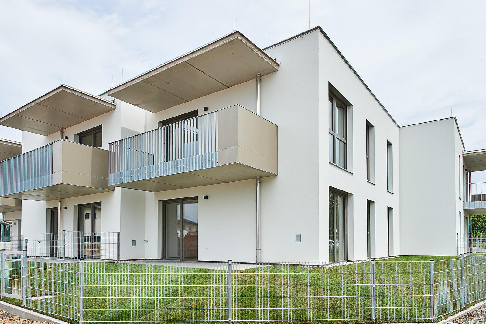 Modernes weißes Wohnhaus mit Balkonen, großen Fenstern und eingezäuntem Garten.