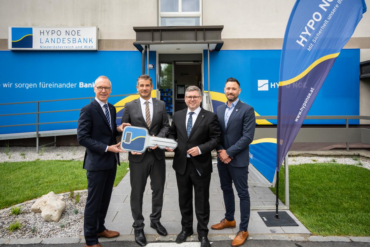 Vier Männer mittleren Alters in Anzügen stehen vor einem Gebäude, dessen Fassade mit dem blau-gelben Logo der HYPO NOE versehen ist.
