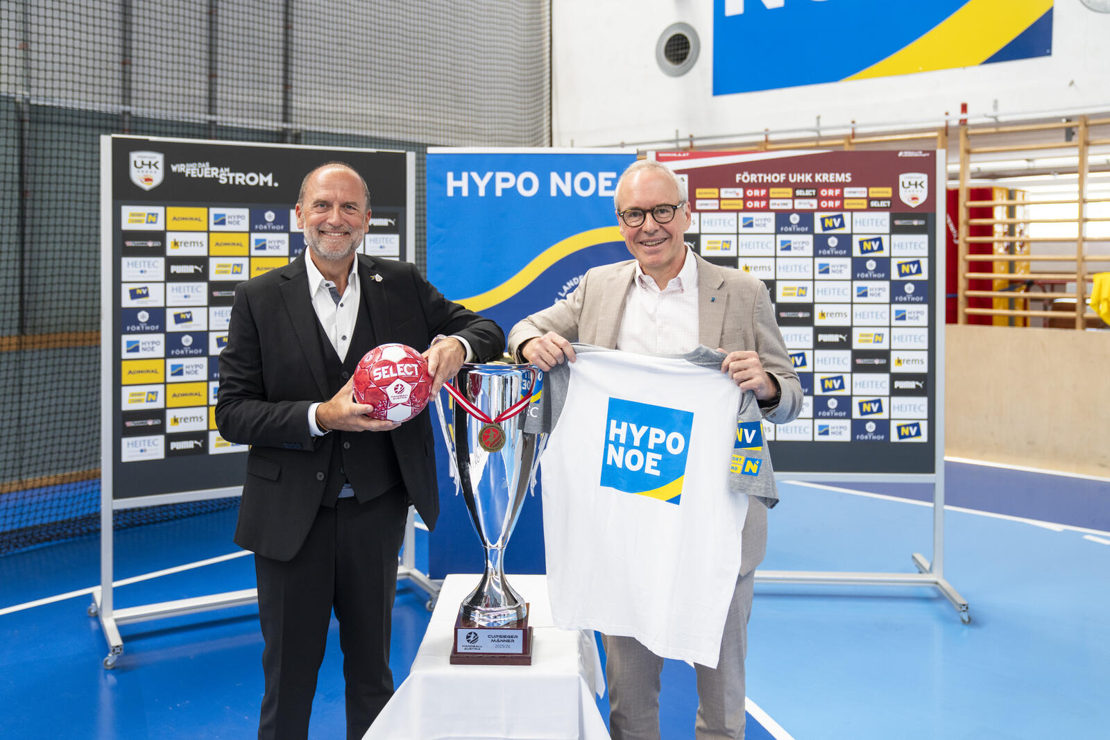 Zwei Personen präsentieren Trikot, Handball und Pokal vor Sponsorenwand.