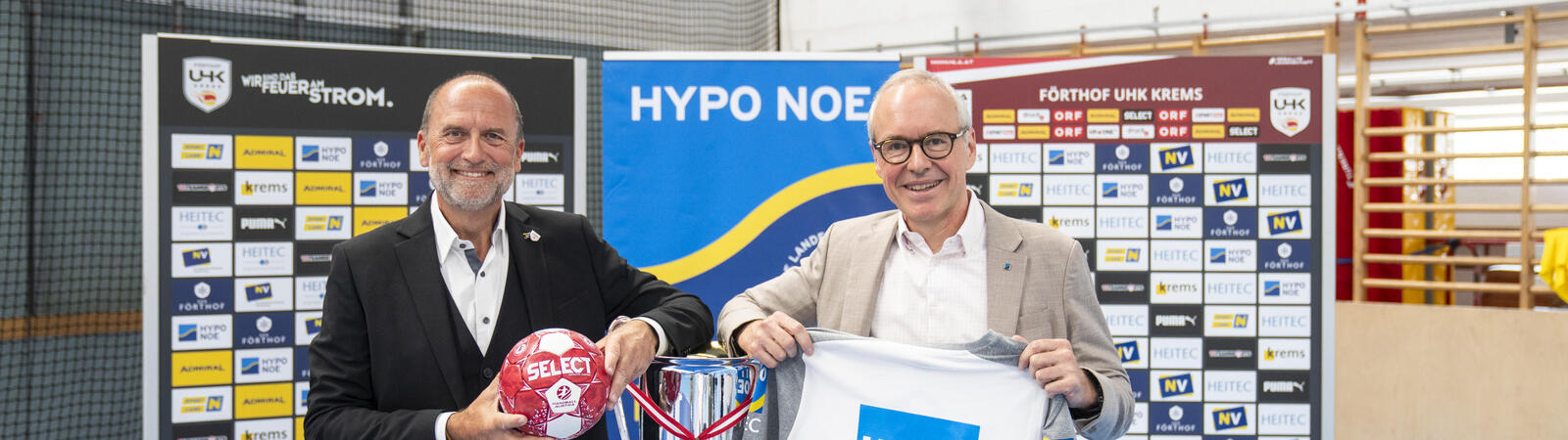 Zwei Personen präsentieren Trikot, Handball und Pokal vor Sponsorenwand.