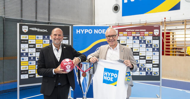 Zwei Personen präsentieren Trikot, Handball und Pokal vor Sponsorenwand.