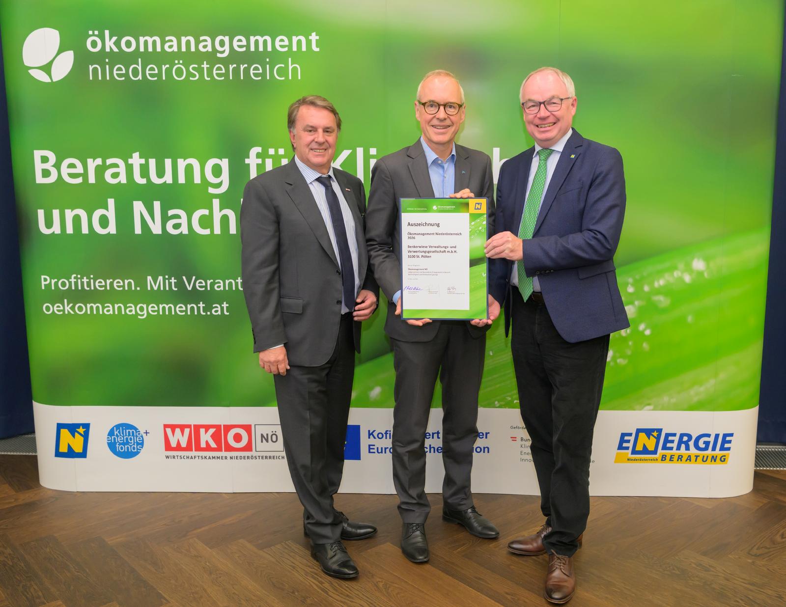 drei Männer in Anzug und Krawatte halten eine Urkunde und stehen vor einer grünen Wand