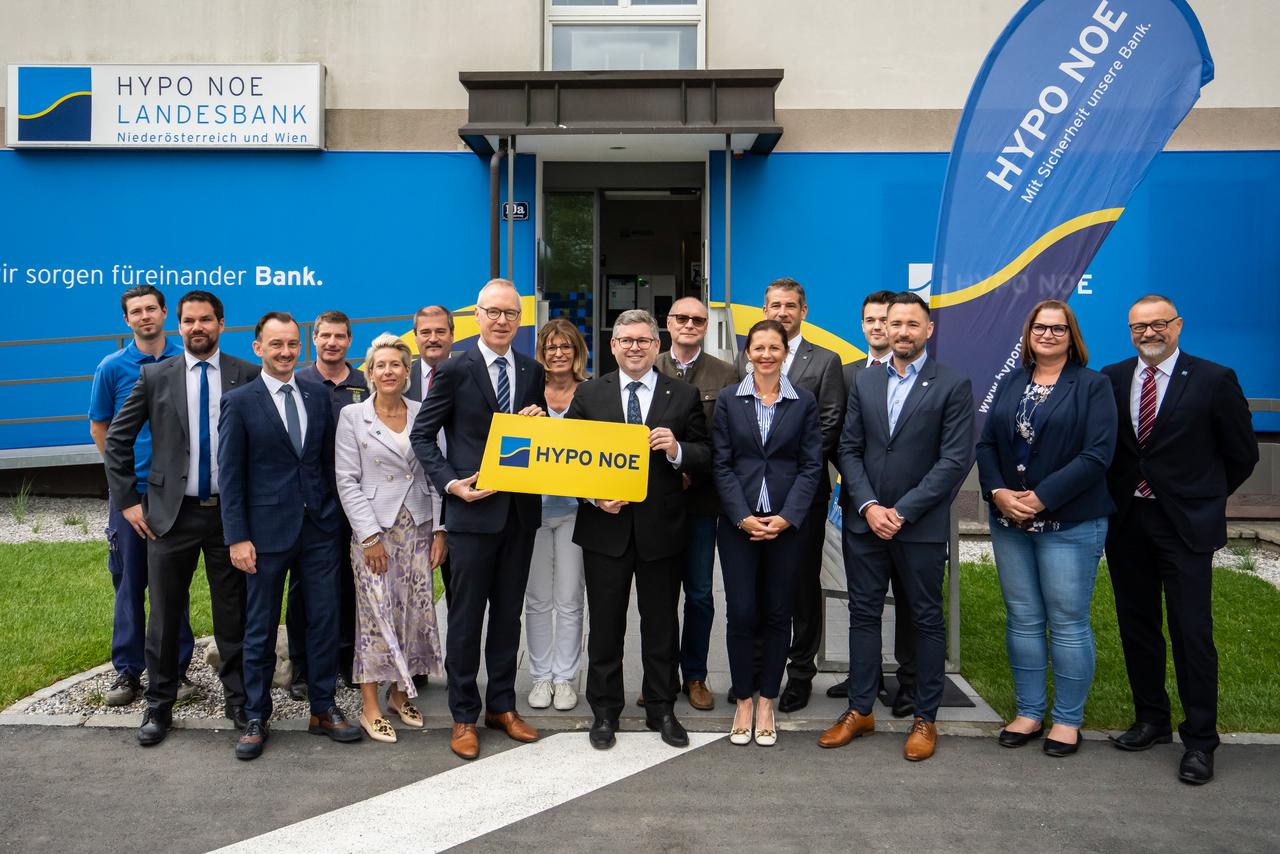 Eine Gruppe von Menschen in Business-Kleidung steht vor einem Gebäude, dessen Fassade mit dem blau-gelben Logo der HYPO NOE versehen ist.