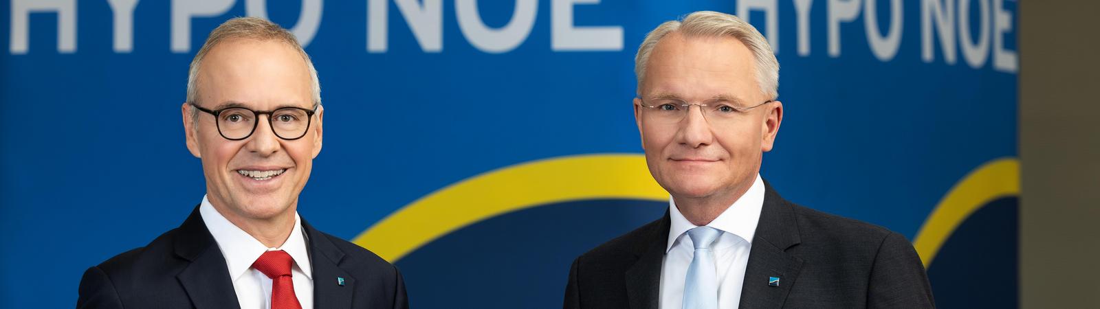 Zwei Männer mittleren Alters im Anzug stehen vor einem dunklen Hintergrund mit Aufschrift "HYPO NOE".