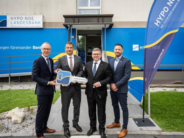 Vier Männer mittleren Alters in Anzügen stehen vor einem Gebäude, dessen Fassade mit dem blau-gelben Logo der HYPO NOE versehen ist.