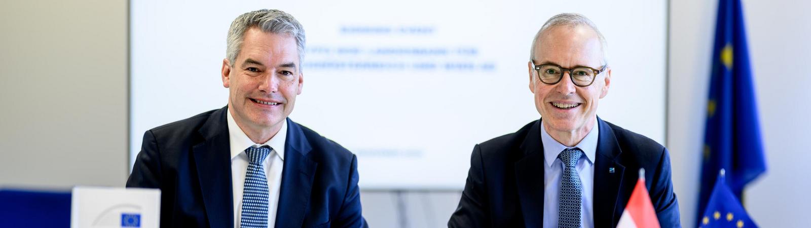 Zwei Männer in Anzug und Krawatte sitzen an einem Tisch und halten Kugelschreiber in der Hand.