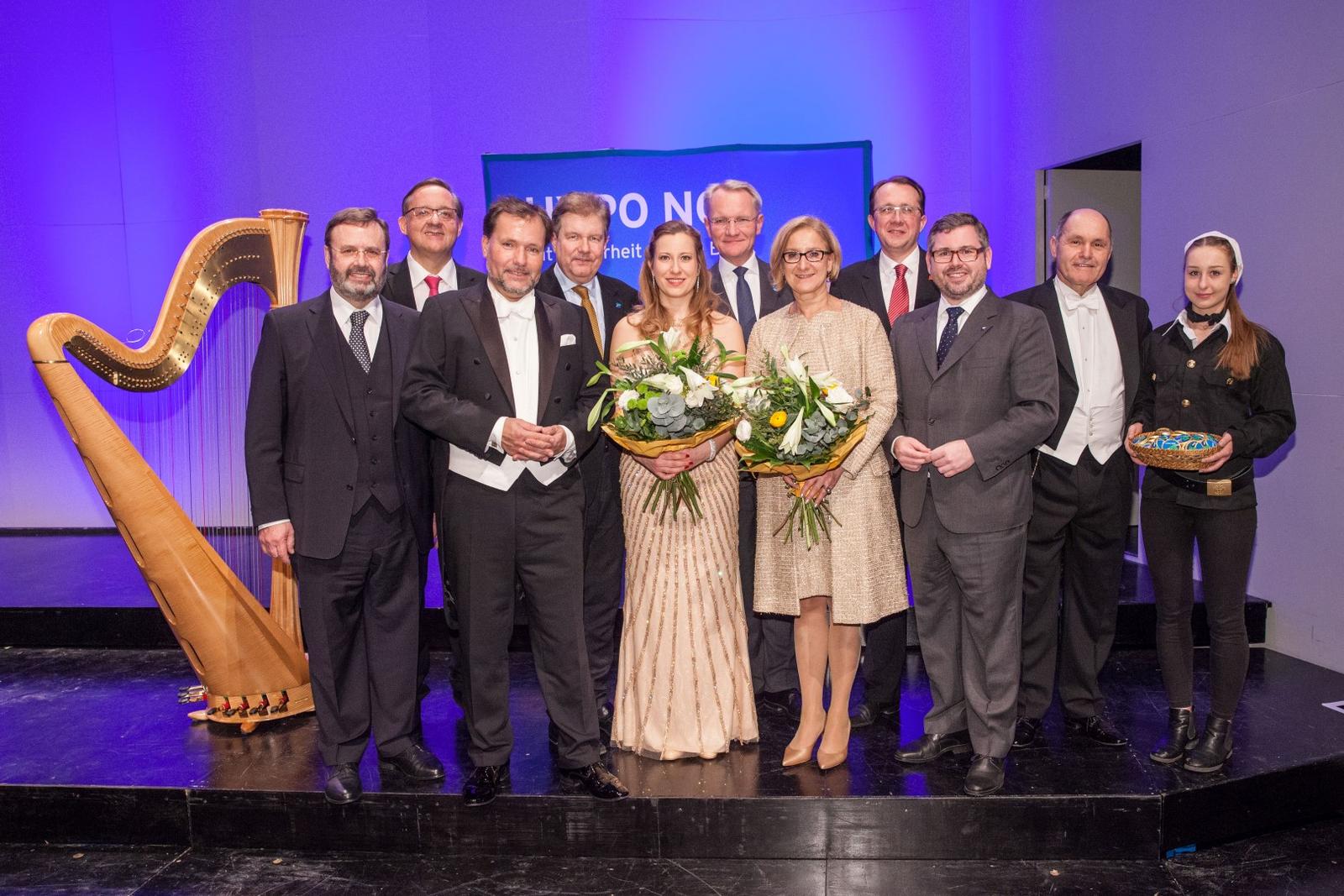 Eine Gruppe von Personen in Abendkleidung stehen auf einer Bühne vor einer Konzertharfe