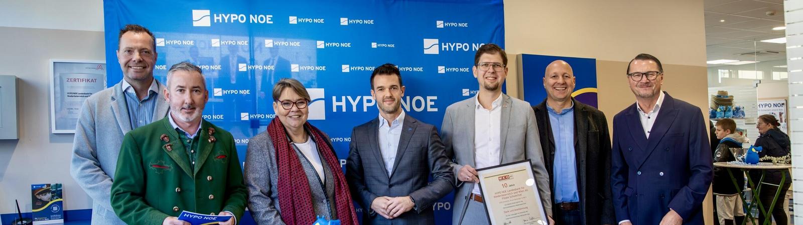 Eine Gruppe von 6 Männern und einer Frau steht vor einer blauen Wand mit Aufschrift "HYPO NOE".