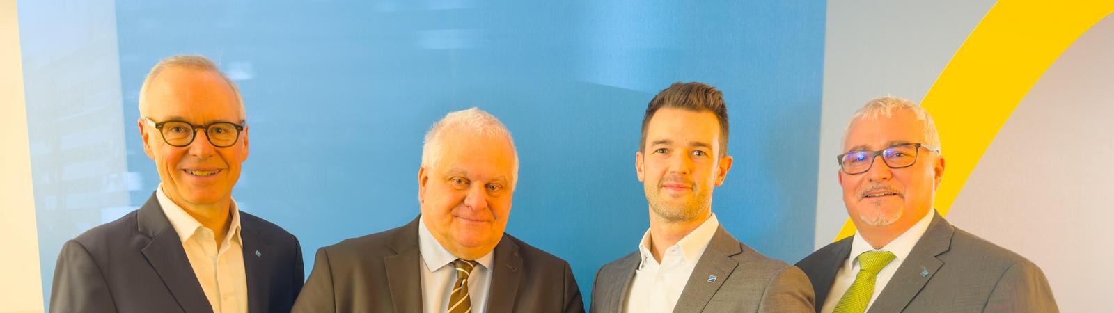 Vier Männer in Anzug stehen nebeneinander, einer hält eine Weinflasche als Abschiedsgeschenk in der Hand.