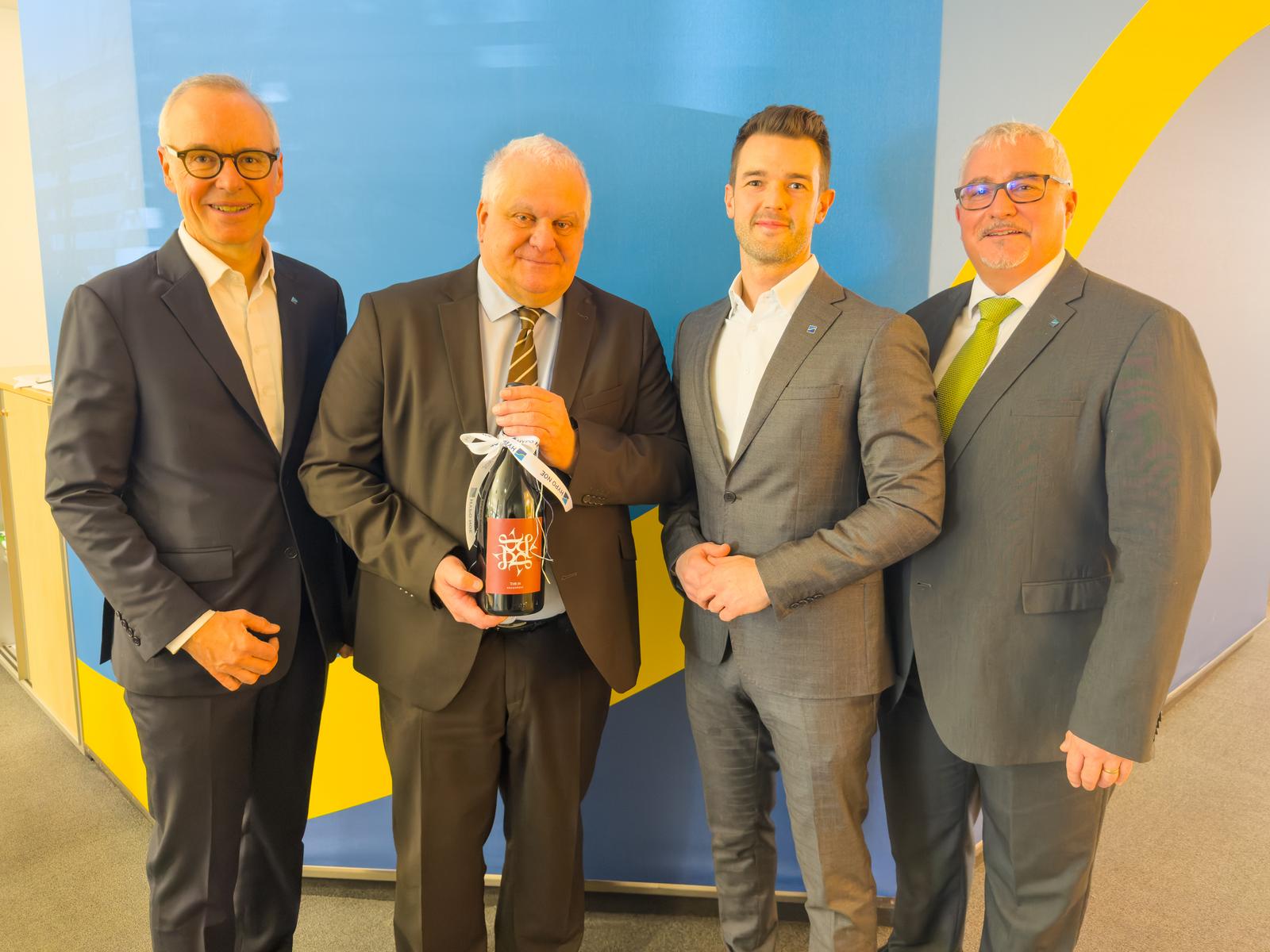 Vier Männer in Anzug stehen nebeneinander, einer hält eine Weinflasche als Abschiedsgeschenk in der Hand.