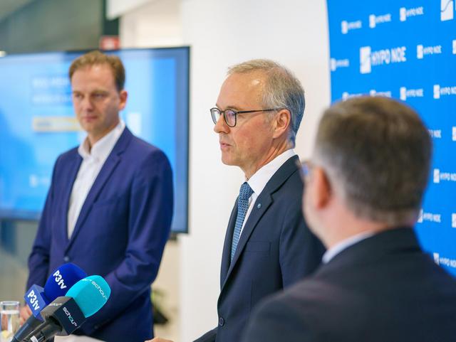 Drei Männer in Anzug und Krawatte an einem Rednerpult mit Mikrofonen.