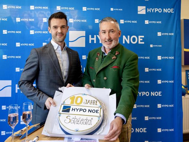 2 Männern und 2 Frauen steht vor einer blauen Wand mit Aufschrift "HYPO NOE" und halten eine Torte in die Kamera.